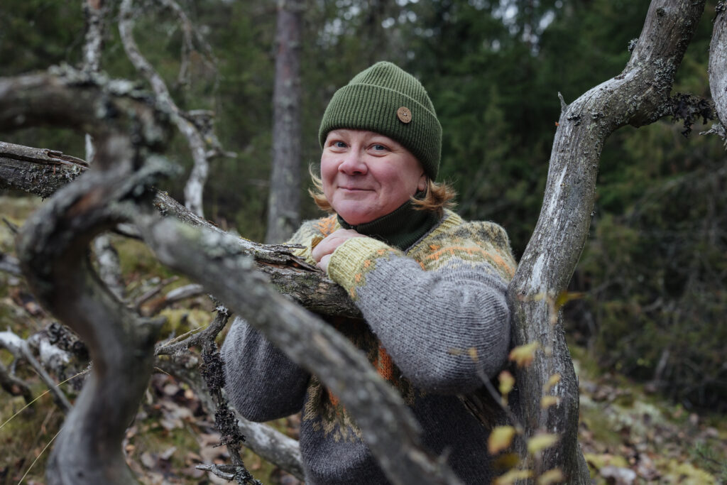 Luontovalmentaja Tarja Jaakkola hymyilee kelottuneiden oksien keskellä päällään harmaa villapaita.